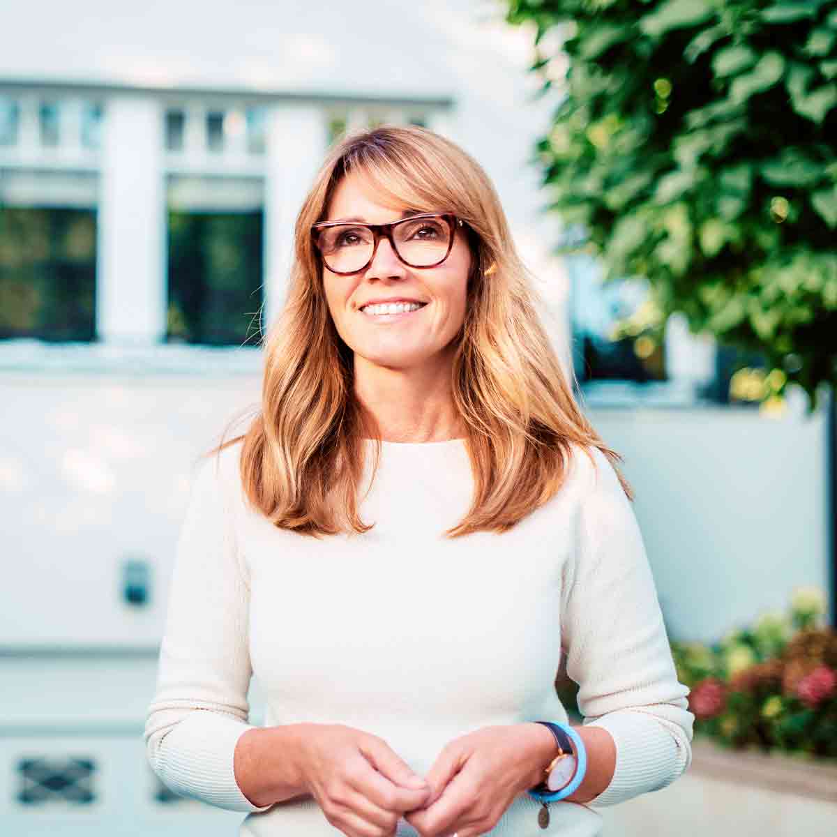 Eastlake Tree review. Smiling woman with blonde hair and glasses standing outdoors wearing a white sweater and watch.