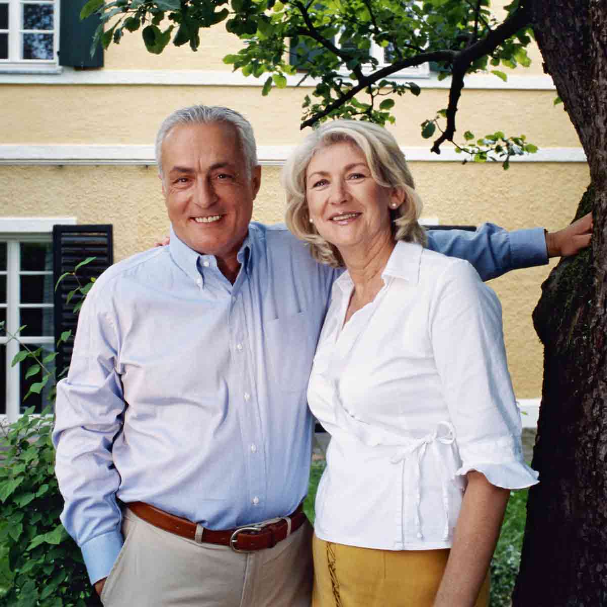 Eastlake Tree Services customer. Smiling elderly couple standing outside near a tree and a yellow house, embracing each other.