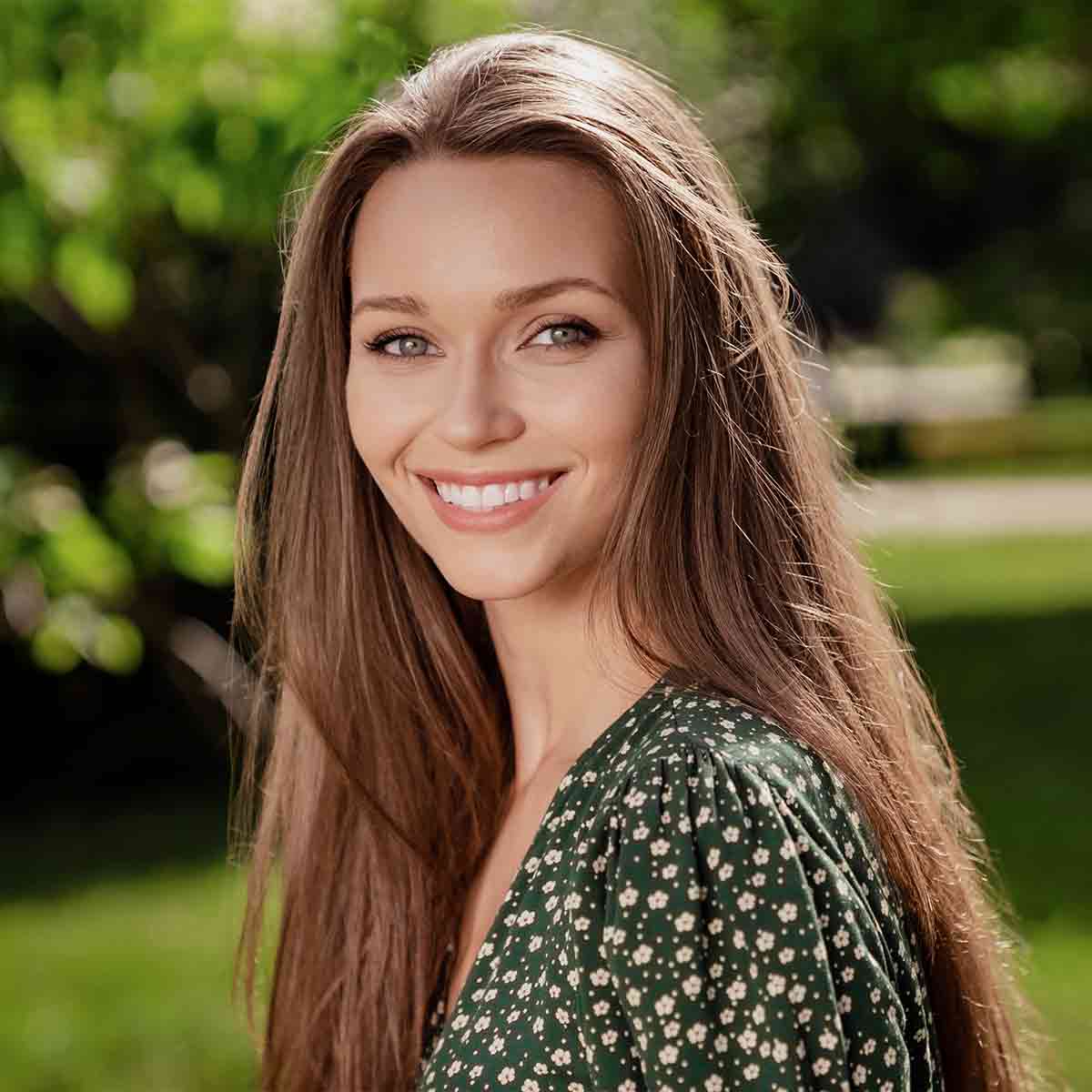 Eastlake Tree Services review. Smiling woman with long brown hair wearing a green floral dress outdoors with blurred greenery in the background.